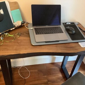 May include: A laptop computer, mouse, and drawing tablet are on a wooden desk. The laptop is silver and has a black keyboard. The mouse is black and the drawing tablet is gray and black. The desk has a live edge and black metal legs.