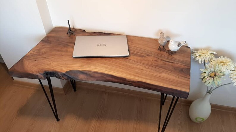 May include: A live edge wooden desk with black hairpin legs. A silver laptop is on the desk, along with decorative birds, a small Eiffel Tower replica, and a vase of flowers. The desk is made of natural wood.
