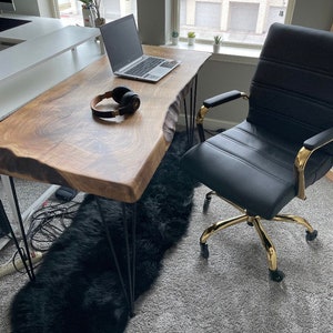 May include: A live edge wooden desk with black hairpin legs. A black leather office chair with gold accents sits to the right. A laptop and headphones rest on the desk. A black faux fur rug is under the desk.