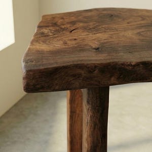 May include: A handcrafted wooden table with a natural, irregular edge. The table is made from dark brown wood, displaying a textured surface. The legs are also wooden and of a similar hue. The table appears robust and is likely handmade, with a height of approximately 45 cm.