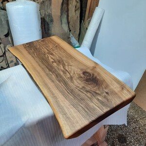 May include: A rectangular wooden serving board with a natural edge. The board is made of light brown wood with dark brown grain patterns. The board is approximately 24 inches long and 12 inches wide. The wood has a smooth, polished finish.