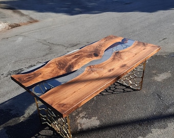 Op de afbeelding: Een houten tafel met een grijze epoxyharsrivier die door het midden stroomt. De tafel heeft een gouden metalen onderstel met een honingraatontwerp. De tafel staat op een grijs asfalt oppervlak.