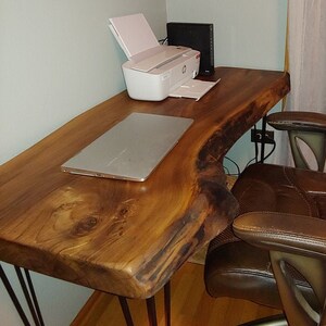 May include: A live edge wooden desk with a natural, warm brown finish. The desk has metal hairpin legs and a laptop on top. A white printer and a black computer tower are also on the desk. A brown leather office chair is next to the desk.