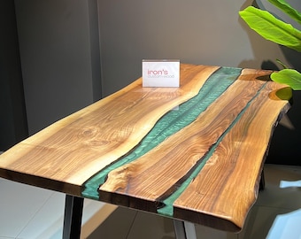 May include: A wooden table with a live edge top and a green epoxy river running through the center. The table has black metal legs in a triangular shape. A small white card with the text "Iron's Custom Wood" is placed on the table.