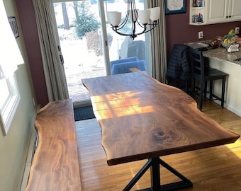 Puede incluir: Una mesa de comedor de nogal de borde vivo con una base de metal negro y un banco de borde vivo a juego. La mesa y el banco están hechos de madera con un acabado natural. La mesa está ubicada en un comedor con vista a un jardín nevado.