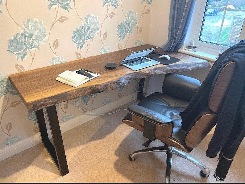 May include: A live edge wooden desk with black metal legs. A laptop, mouse, notebook, and pen sit on the desk. A black leather office chair is in front of the desk. The desk is in a room with floral wallpaper.