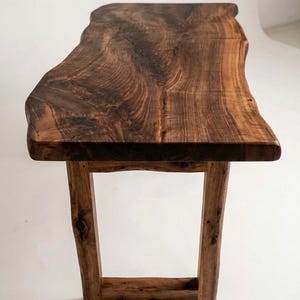 May include: A wooden bench with a live edge top, showcasing a rich, dark brown wood grain. The bench is supported by four wooden legs. The design highlights the natural beauty of the wood, making it a unique piece of furniture. The bench is approximately 120 cm long.