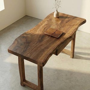 May include: A wooden desk with a natural edge and a warm brown finish. A small leather-bound journal rests on the desktop, along with a small vase holding a few sprigs. The desk has a minimalist design with sturdy, square legs.
