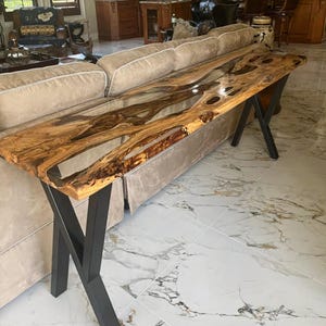 Puede incluir: Una mesa larga de madera con una tapa de resina epoxi transparente. La mesa tiene un patrón de veta de madera natural y está soportada por una base de metal negro en forma de X.