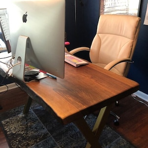 May include: A wooden desk with a live edge top and gold metal legs. A computer monitor and keyboard are on the desk. A tan leather office chair is in the background.