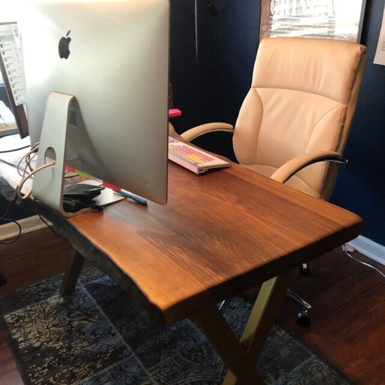 May include: A wooden desk with a live edge top and gold metal legs. A computer monitor and keyboard are on the desk. A tan leather office chair is in the background.