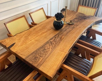 Pode incluir: Uma grande mesa de jantar de madeira com um design de borda viva. A mesa é feita de madeira escura com um acabamento natural. É cercada por seis cadeiras com encostos de vime trançado e assentos de couro escuro.