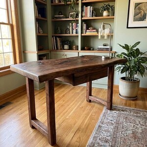 May include: A dark brown wooden desk with a single drawer, set against a backdrop of built-in bookshelves and a potted plant. The desk's solid construction and natural wood grain are prominent features. The room has a rug.