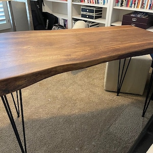 May include: A live edge wooden table with a natural, irregular edge. The tabletop is a rich brown color and is supported by black metal hairpin legs. The table measures approximately 72 inches long.