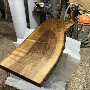 May include: A large, dark brown wooden slab with a live edge. The slab is smooth and polished, with a natural grain pattern. It is sitting on a white surface.