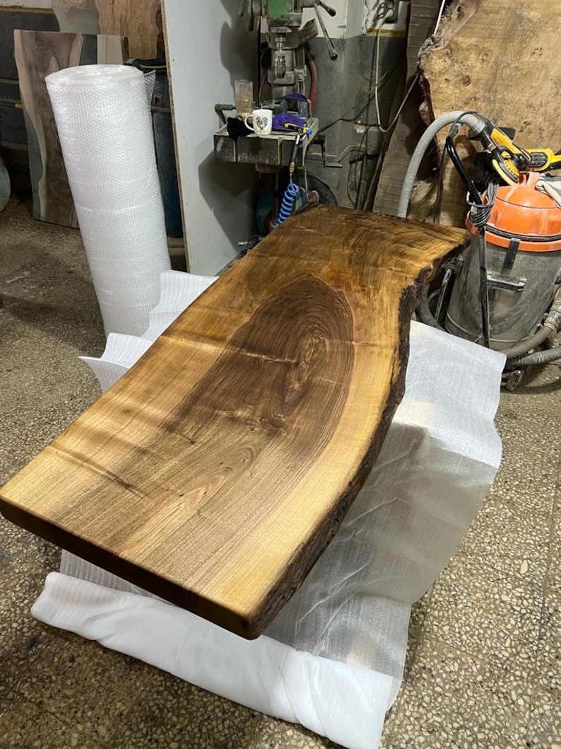 May include: A large, dark brown wooden slab with a live edge. The slab is smooth and polished, with a natural grain pattern. It is sitting on a white surface.