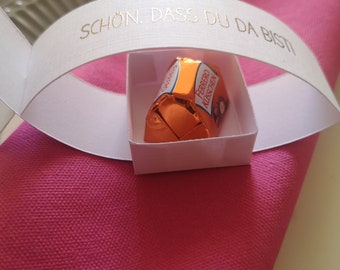 Op de afbeelding: Een klein vierkant wit papieren doosje met daarin een Ferrero Küsschen chocolaatje verpakt in oranje folie. Een witte papieren band boogt over het doosje met de goudkleurige tekst SCHÖN. DASS DU DA BIST, rustend op een felroze servet.