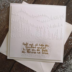 Könnte beinhalten: Eine weiße Grußkarte mit geprägtem Waldmotiv und goldenem Text. Die Karte liegt auf einem weißen Umschlag. Der Text lautet: "Denkt euch, ich habe das Christkind gesehent ty Kam quy dem Walde, day Mitzchen voll Schies."