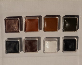 May include: A set of eight watercolor paint pans in a white plastic tray. The pans are arranged in two rows of four. The colors are brown, orange, tan, green, black, brown, white, and black.