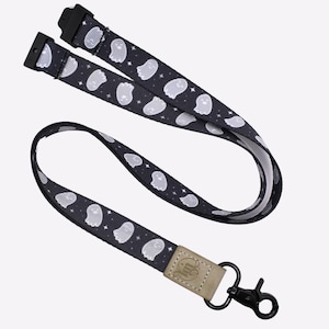 Puede incluir: Lanyard negro con un patrón de fantasmas blancos y un clip metálico negro. El lanyard tiene un parche de cuero marrón con un logotipo.