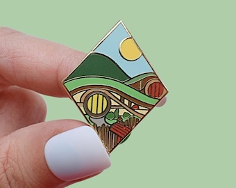 Könnte beinhalten: Ein rautenförmiger Emaille-Pin mit einer Landschaftsdarstellung. Der Pin zeigt grüne Hügel, einen blauen Himmel mit einer gelben Sonne und ein Hobbit-Loch mit einer runden Tür und einem Holzzaun. Der Pin hat einen goldenen Rand.