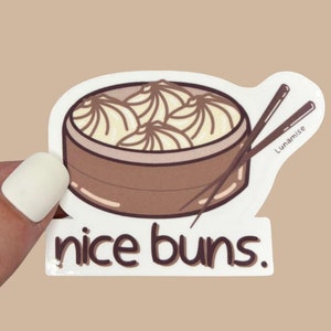Könnte beinhalten: Ein Aufkleber mit einer Cartoon-Illustration eines Bambusdämpferkorbs, gefüllt mit Knödeln, mit Essstäbchen und dem Text "nice buns". Der Aufkleber ist in Braun- und Cremetönen gehalten.