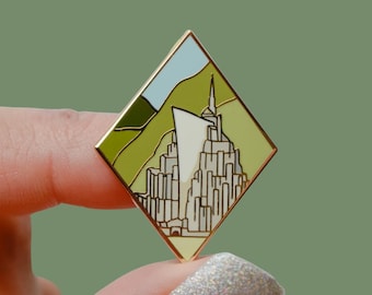 Könnte beinhalten: Ein goldfarbener Emaille-Pin in Diamantenform. Der Pin zeigt eine stilisierte Illustration eines weißen Bauwerks mit hohem Turm vor einer grünen und blauen Landschaft. Der Pin wird zwischen Fingern vor grünem Hintergrund gehalten.