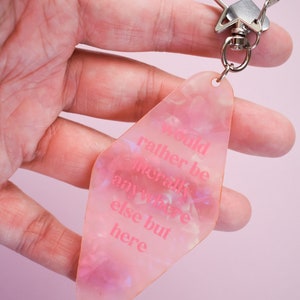 Könnte beinhalten: Rosa diamantförmiger Schlüsselanhänger mit dem Text "would rather be literally anywhere else but here" in passender rosa Schrift. Der Schlüsselanhänger hat einen silbernen Verschluss und Ring.