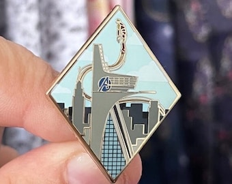 Könnte beinhalten: Eine goldfarbene Emaille-Anstecknadel in Form eines Diamanten. Die Anstecknadel zeigt eine Stadtlandschaft mit einem hohen Gebäude und einer fliegenden Figur im Himmel. Das Gebäude hat ein großes "A"-Logo an der Seite.