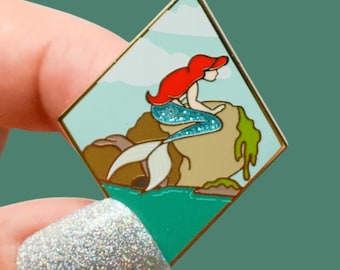 Könnte beinhalten: Ein rautenförmiger Emaille-Pin mit einer Meerjungfrau mit roten Haaren und blauem Schwanz, die auf einem Felsen sitzt. Der Pin hat einen goldenen Rand und einen türkisfarbenen Hintergrund mit einem grünen und braunen Felsen. Die Meerjungfrau ist eine Cartoon-Illustration.