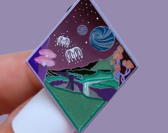 Könnte beinhalten: Ein rautenförmiger Emaille-Pin mit einer Fantasy-Landschaft. Das Design zeigt einen Sternenhimmel mit Planeten, schwebenden quallenartigen Kreaturen und bunten Pilzen. Der Pin hat ein grünes, lila und blaues Farbschema.
