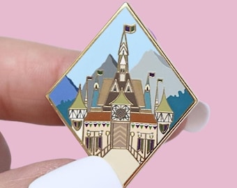 Könnte beinhalten: Ein goldfarbener Emaille-Pin in Diamantform mit einer detaillierten Illustration eines Schlosses mit Bergkulisse. Das Schloss hat mehrere Türme, Flaggen und eine Brücke. Der Pin wird zwischen Fingern vor einem rosa Hintergrund gehalten.