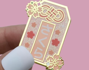 May include: A gold-colored enamel pin with a pink and white design. The pin features the Japanese word "Sakura" in white, cherry blossom accents, and a decorative knot. The pin is held against a pink background.