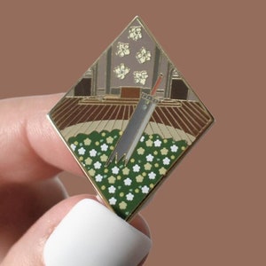 Puede incluir: Un pin de esmalte de color dorado en forma de diamante con un diseño floral verde y blanco. Una espada plateada está sobre las flores. El pin tiene un fondo marrón y dorado con un edificio blanco en el fondo.