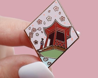 Könnte beinhalten: Ein rautenförmiger Emaille-Pin mit goldenem Rand, der eine japanische Pagoden-Szene darstellt. Der Pin zeigt eine rote Pagode, eine Person in traditioneller Kleidung, Kirschblüten und eine grüne und blaue Landschaft vor einem rosa Hintergrund.
