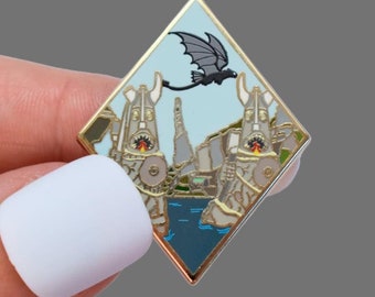 May include: A diamond-shaped enamel pin featuring a fantasy landscape with a flying dragon. The pin has a gold border and a light blue sky background. The landscape includes two tall structures, water, and mountains.