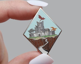 May include: A diamond-shaped enamel pin featuring a fantasy castle scene with a winding road and flying dragons. The pin has a light blue sky, a brown cliff, and a silver border. The dragons are red and orange.