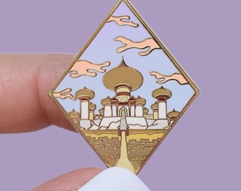 May include: A gold-colored enamel pin in a diamond shape, featuring a cartoon illustration of a palace with a golden dome, set against a blue sky with pink clouds. The pin has a gold border and is held against a purple background.
