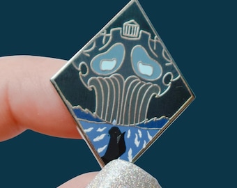 Könnte beinhalten: Ein rautenförmiger Emaille-Pin mit dunkelblauem Hintergrund. Der Pin zeigt ein stilisiertes Design mit hellblauen und silbernen Akzenten, darunter ein Wasserfall und eine schwarze Figur. Der Pin wird zwischen zwei Fingern gehalten.