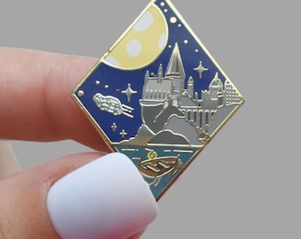 Könnte beinhalten: Ein rautenförmiger Emaille-Pin mit einer Nachtszene mit einer Burg, einem Mond, Sternen und einem Boot. Der Pin hat einen goldenen Rand und einen blauen Hintergrund. Die Burg ist grau mit weißen Akzenten. Der Mond ist gelb mit weißen Punkten.