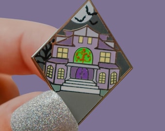 Könnte beinhalten: Ein rautenförmiger Emaille-Pin mit einem lila Geisterhaus-Design. Das Haus hat gelbe Fenster, ein grünes Buntglasfenster und eine lila Tür. Ein schwarzer Baum, Fledermäuse und ein Mond sind im Hintergrund.