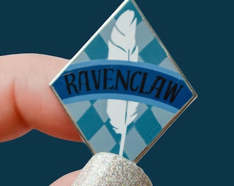 Könnte beinhalten: Ein rautenförmiger Ravenclaw-Anstecker mit silbernem Rand. Der Anstecker zeigt ein blau-weißes Rautenmuster, eine weiße Feder und das Wort "RAVENCLAW" in blauen Buchstaben auf einem blauen Banner.