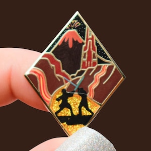 Puede incluir: Un pin de esmalte en forma de diamante que representa una escena con dos figuras luchando con sables de luz. El fondo incluye un volcán, una torre y un cielo estrellado. El pin tiene un borde dorado y una base dorada brillante.