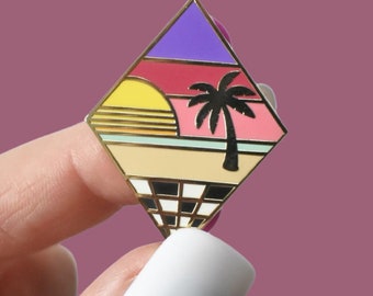 May include: A diamond-shaped enamel pin featuring a sunset scene with a palm tree. The pin has a gold border and a checkered pattern at the bottom. The sunset includes yellow, pink, and purple hues. The pin is held against a purple background.