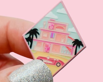 Könnte beinhalten: Ein pinkfarbener Emaille-Pin mit einem zweistöckigen rosa Haus mit einem roten Cabrio auf einer gelben Straße. Der Pin hat einen hellblauen Himmel mit weißen Wolken, schwarzen Palmen und einen pinkfarbenen Rand.