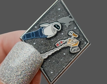 Könnte beinhalten: Ein rautenförmiger Emaille-Pin mit den Charakteren Wall-E und Eve aus dem Animationsfilm. Der Pin hat einen silbernen Rand und einen sternenübersäten Hintergrund. Eve ist weiß und blau, während Wall-E orange und braun ist.