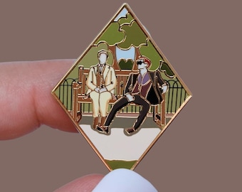 May include: Diamond-shaped enamel pin depicting two figures on a park bench. The pin has a gold border, with a green and blue background. One figure wears a suit, the other wears sunglasses and a black suit.