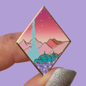 Puede incluir: Un pin de esmalte en forma de diamante con borde dorado. El pin presenta un paisaje con un cielo rosa y morado, montañas, un castillo azul claro y cúpulas turquesas. La parte inferior del pin tiene un patrón de estrellas moradas y blancas.