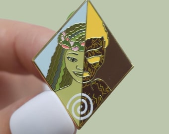 Könnte beinhalten: Ein rautenförmiger Emaille-Anstecker mit goldenem Rand, der ein geteiltes Design aufweist. Eine Seite zeigt eine Frau mit Blumenkranz, die andere eine strukturierte, dunkle Figur. Der Anstecker hat einen goldenen Rand und einen grünen Nadelrücken.