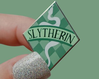 Könnte beinhalten: Ein rautenförmiger Slytherin-Anstecker in Grün und Weiß. Der Anstecker zeigt eine weiße Schlange und das Wort "Slytherin" auf einem grünen Banner. Der Hintergrund hat ein grün-weißes Karomuster.